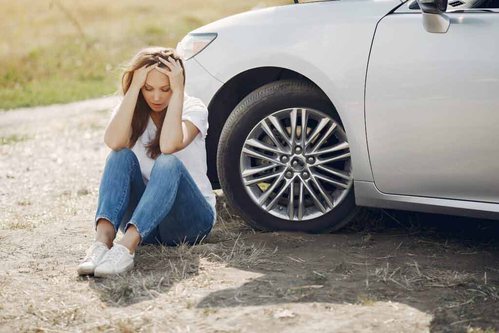 A distressed young woman sitting on the ground in front of her vehicle after an accident.